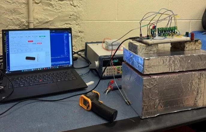 The full bench on a lab table — laptop running the logging dashboard, BK Precision 8601 electronic load, and a foil-wrapped foam enclosure with granite weights stacked on the lid and the ESP32 thermocouple breadboard sitting on top.
