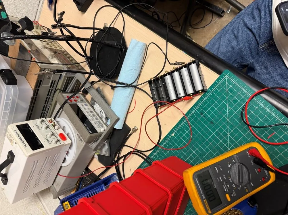 Bench top with four 21700 cells in a black holder wired to a benchtop DC power supply, alongside a multimeter reading zero and a cutting mat.