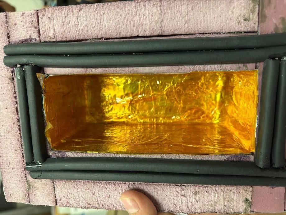 The empty thermal enclosure held up to the camera — pink extruded foam body, black rubber gasket cord around the rim, and a gold Kapton film lining the inner cavity.