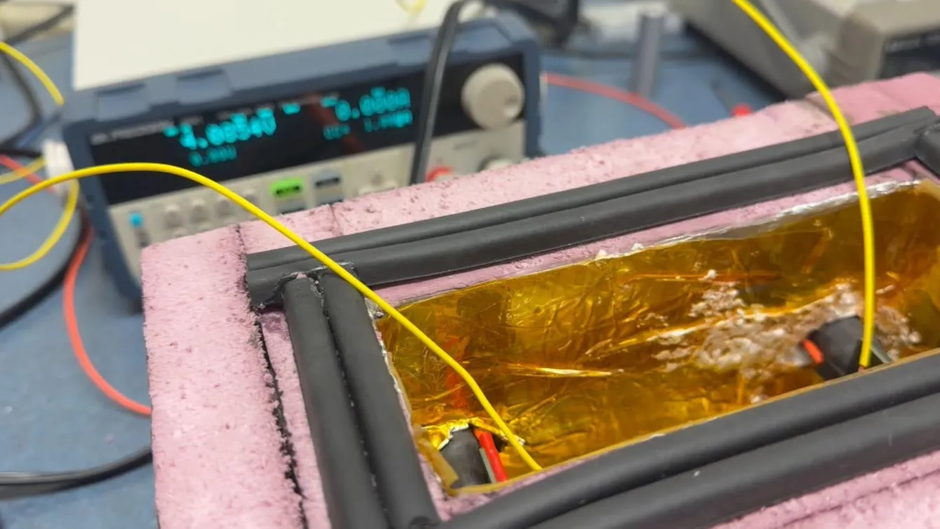 A cell inside the Kapton-lined cavity of the pink foam enclosure, yellow type-K thermocouple leads running in over the black gasket cord, with the electronic load's display lit in the background.