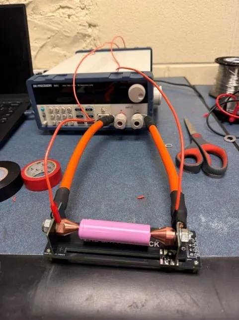 Close view of the test fixture wired to a BK Precision 8601 load — thick orange force leads to the copper cone contacts, thin red sense wires tapping near the same lugs.