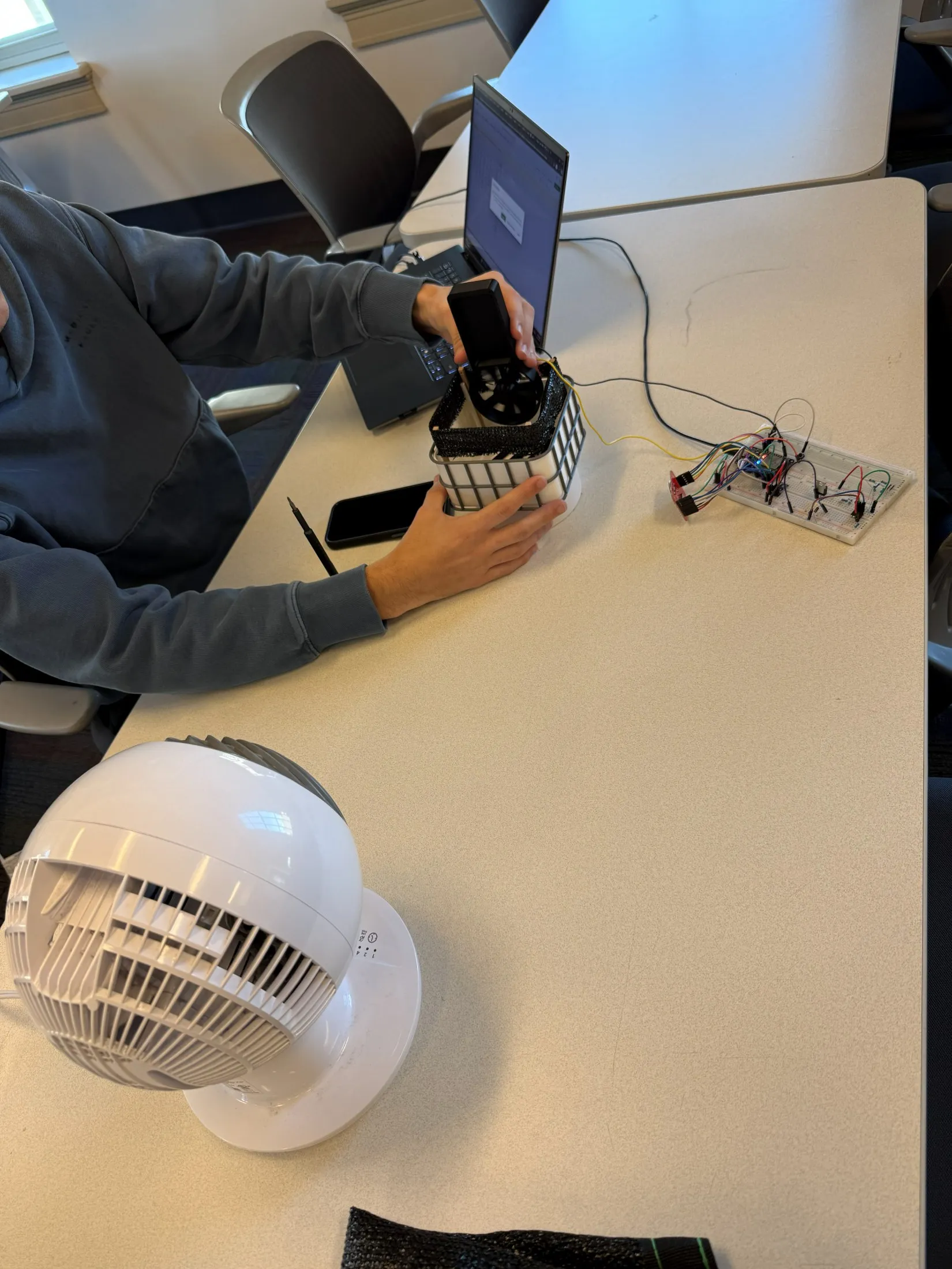 The wind test in progress — a fan in the foreground aimed at the mesh-wrapped microfarm, the anemometer held over it, with a breadboard of electronics alongside.